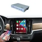Center console of a car with a large tablet-style screen in the dash; a small wireless car adapter sits above, while a hand holds a smartphone nearby.