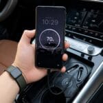 Phone shows 70% battery while charging wirelessly in a car's center console, held by a person wearing a smartwatch.