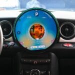 Circular digital dashboard displaying music player with a portrait artwork in the center, car interior around it. More concise: Infotainment screen showing music progress inside a round display in a car dashboard.